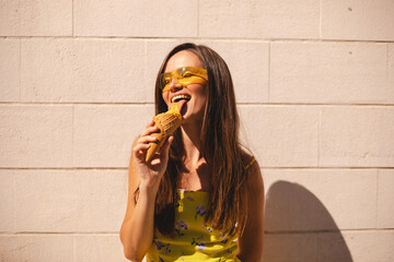 Close up happy young woman with delicious yellow ice cream in waffle cone outdoors. Girl wear yellow sunglasses in summer, spring or fall sunny day. Brunette female lick ice cream and laughing.