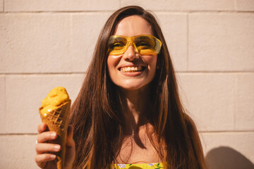 Close up happy young woman with delicious yellow ice cream in waffle cone outdoors. Girl wear yellow sunglasses and dress in summer, spring or fall sunny day. Brunette female laughing.