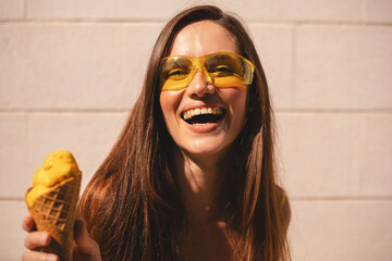 Close up happy young woman with delicious yellow ice cream in waffle cone outdoors. Girl wear yellow sunglasses and dress in summer, spring or fall sunny day. Brunette female laughing.