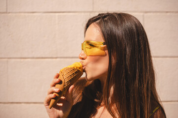 Close up happy young woman with delicious yellow ice cream in waffle cone outdoors. Girl wear yellow sunglasses in summer, spring or fall sunny day. Brunette female lick ice cream and laughing.
