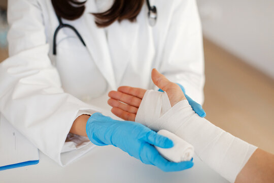 First aid in wrist injuries. Traumatologist taking care of female patient, woman doctor bandaging hand, sitting at table in clinic office