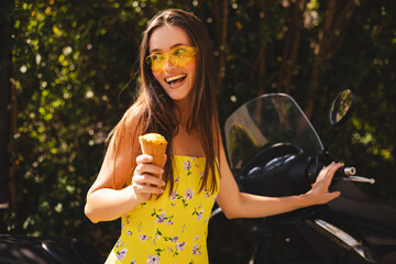Happy laughing brunette long hair woman standing near her motor bike moped, hold ice cream and smiling. Woman wear bright yellow dress and sunglasses, summer mood.