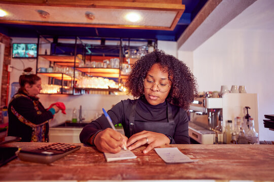 Concentrated waitress writing down orders in a notepad at the counter of a cozy bar, coworker cleaning in the background