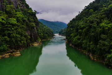 Fototapeta premium Wahrew river, a green river flows between steep, forested mountains under a cloudy sky.