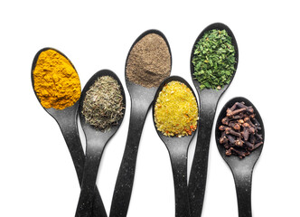 Top view of six black spoons filled with colorful spices and herbs on a white background