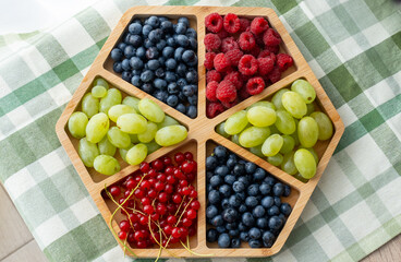 Assorted fresh berries and grapes arranged in hexagonal wooden serving tray on green checkered tablecloth. Colorful mix of blueberries, raspberries, red currants and green grapes for healthy eating.