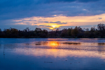 sunset over a calm river with fiery sky reflection in water, autumn landscape with tree silhouettes on the horizon
