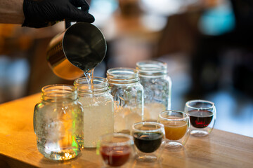 Gloved hand pouring liquid into mason jar with assorted drinks on wooden table, front view, isolated in cafe setting.