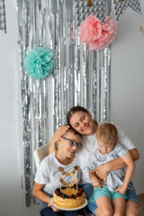 Loving mother with two sons enjoying homemade birthday cake with fresh fruit decoration and sparkly backdrop. Warm family moment during home celebration with metallic streamers and golden cake topper.