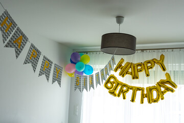 Happy Birthday golden foil letter balloons and colorful party decorations hanging in bright modern living room with bunting flags. Festive home celebration setup for birthday party or anniversary.