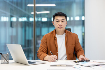 An Asian businessman in a modern office setting, working at his desk. He's focused, looking directly at the camera.