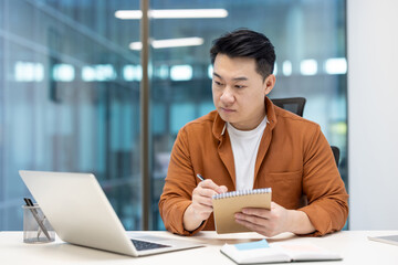 Fototapeta premium An Asian man sits at his desk, concentrating while taking notes and using his laptop in a modern office setting.