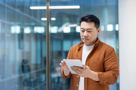 An Asian man in an office setting using a tablet. The background has glass windows. - Powered by Adobe
