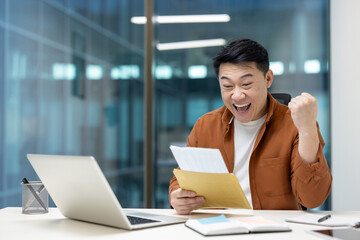 An excited Asian man celebrates with a raised fist after reading good news from a letter. He's...