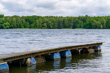 Lakeside landscape with a tilted wooden pier floating on plastic barrels. Calm water, dense forest in the background, and a cloudy sky above.