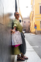elegant mature woman with colorful shopping bags looking into camera leaning with her back against a black wall