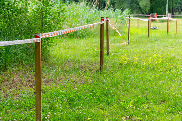 Wooden stakes connected by red and white warning tape on a grassy field. The tape marks a restricted or designated area