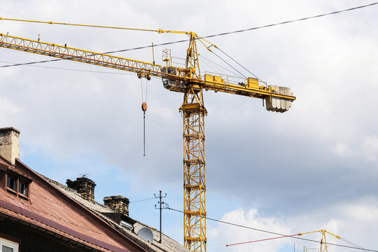 Yellow construction crane above rooftops. Industrial equipment on building site. Urban development project. Cloudy blue sky background. Residential housing renovation. Heavy machinery outdoors.