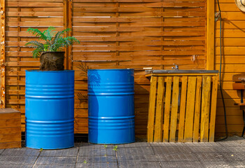 Two blue metal barrels in front of a wooden wall, one decorated with a plant in a jute sack. Next...