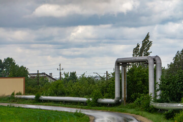 Fototapeta premium Heating pipes running above a bend in an asphalt road, partially surrounded by greenery. In the background, buildings and power lines under a cloudy, slightly stormy sky.