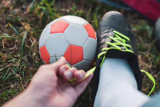 A close-up of a soccer scene outdoors, focusing on a vibrant orange-and-white soccer ball and a black soccer shoe with bright green laces. The grassy field and natural light add a summery, active vibe - Powered by Adobe