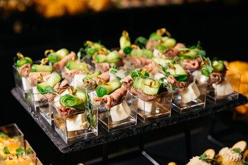 Mini beef and cucumber appetizers in cups. Gourmet hors d'oeuvres served in small clear cups, featuring rolled beef slices, cucumber ribbons, and cheese cubes.