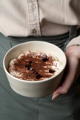 A person holds a bowl of dessert with creamy mascarpone layers, sprinkled with cocoa powder and topped with chocolate pieces, showcasing artistry in a modern kitchen