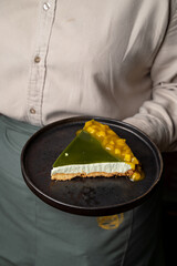 A friendly server presents a slice of dessert, showcasing layers of vibrant green jello atop creamy filling and a golden mango topping on a flaky crust, creating an inviting treat for guests