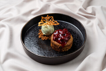A scrumptious dessert is expertly presented on a black plate, showcasing a chocolate tart topped with vibrant cherry compote and a mint ice cream scoop alongside crispy cookie crumbles