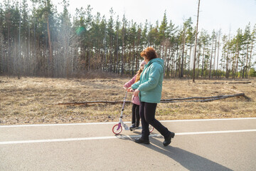Fototapeta premium Little girl riding scooter while walking with her grandmother in the forest on a sunny day