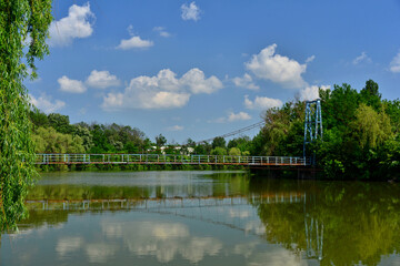 The lake of the river Teleajen as it flows through the Bucov park with the pedestrian suspension bridge and cumulus clouds in the blue sky