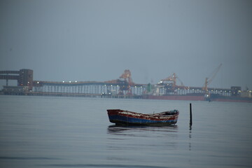 Naklejka premium Small, weathered rowboat sitting in calm water, showing signs of age and wear. In the background, a large industrial port.