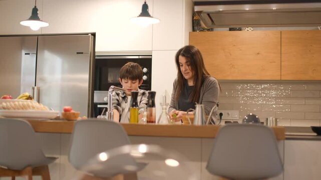 Wide frontal shot of a mother and her son cooking side by side at a modern kitchen island. The boy helps by washing vegetables, sharing household tasks in natural light. Horizontal, 4K