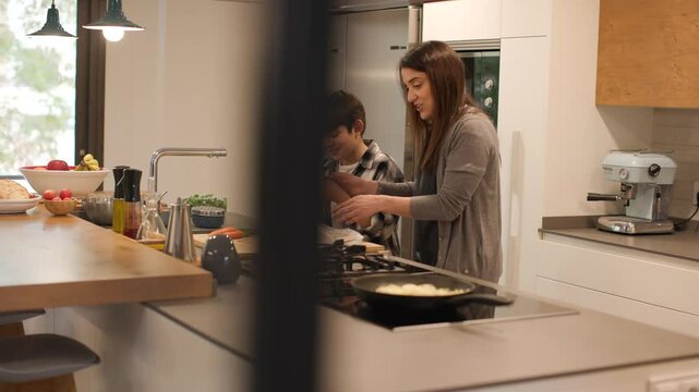Wide distant shot seen through the kitchen door: a bright, modern kitchen where a mother and her son cook together at the island. She smiles while teaching him how to cut vegetables safely. Horizontal
