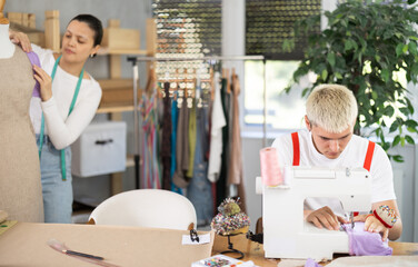 Attentive young tailor using sewing-machine while Asian girl working with mannequin in sewing...