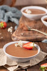 Individual servings of smooth chocolate mousse in white bowls, garnished with fresh strawberries and tiny white flowers, arranged on a rustic wooden board and parchment paper