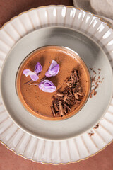 A very close overhead shot captures creamy chocolate mousse in a glass, perfectly garnished with chocolate shavings and purple flowers on decorative plates.