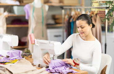 Positive young female dressmaker working with sewing machine in tailor's workshop