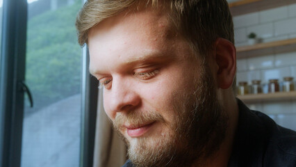 Bearded man with a gentle smile looks down near a window, captured in soft natural light, peaceful...