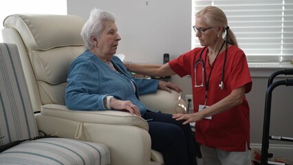 Obraz premium A caregiver in a red uniform talks compassionately to an elderly woman in a blue sweater while seated in a cozy living room with natural light.
