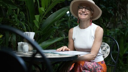 Fototapeta premium A mature woman sits at a cafe table, reading a menu. Surrounded by plants, she immerses herself in work during her vacation, showcasing a digital nomad lifestyle.