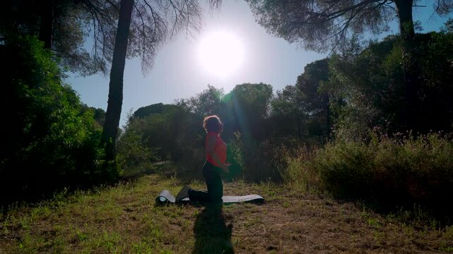 Mujer haciendo yoga en el bosque con el sol de fondo a contraluz y los reflejos de los rayos de luz que la iluminan. Realiza ejercicios de respiraci&oacute;n de rodillas y sentada.