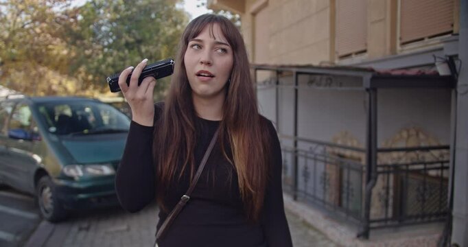 A young woman stands on a city sidewalk making a variety of facial expressions while holding her phone to her ear. She listens intently as she talks.