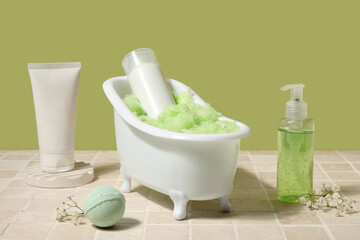Composition with mini bathtub, bottles of cosmetic products and bomb on tile table near green wall