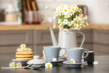 Watering can with flowers, cups of natural chamomile tea and cookies on black table in kitchen