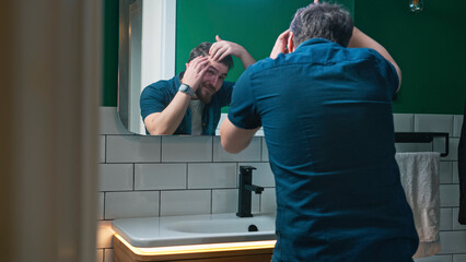 A man stands in front of a bathroom mirror, looking worriedly at his hair, checking for signs of hair loss.