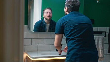 A man leans over the sink, splashing water onto his face with both hands as he washes up, looking focused and intent on his morning routine. He rubs his face to refresh and wake up.
