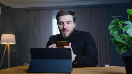 A man sits in a modern room holding phone in one hand while looking at a tablet on the desk. Focused man reviewing documents or working on administrative tasks