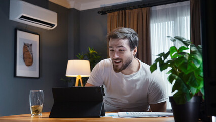 A man in a casual white shirt participates in an online video call meeting at home. He holds up paper reports to show to the camera while actively talking and explaining.