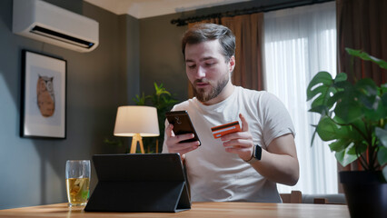 A man holds a smartphone and a credit card, making an online transaction in a living room. Digital shopping or banking in a modern home